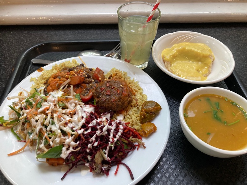 A tray with: a drink, a plate with two curries, rice, beetroot salad, and bean sprout salad, a bowl of soup, and a bowl of custard